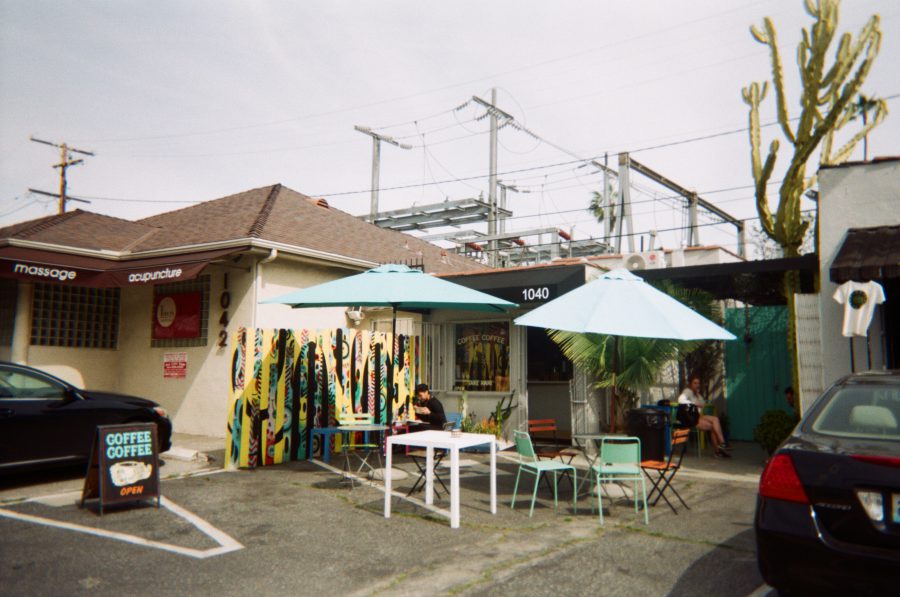 The exterior of Coffee Coffee in West Hollywood, featuring vibrant hand-painted wooden panels and light blue umbrellas over outdoor seating. A couple of patrons relax at colorful chairs and tables, while the signage for acupuncture and massage next door adds a local flair.