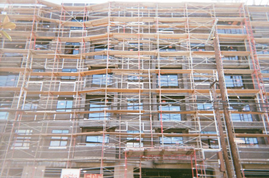 A large building under construction with an intricate network of scaffolding covering the facade. The bright sunlight highlights the wooden planks and metal supports against the modern design of the structure, with glass windows visible between the scaffolding.