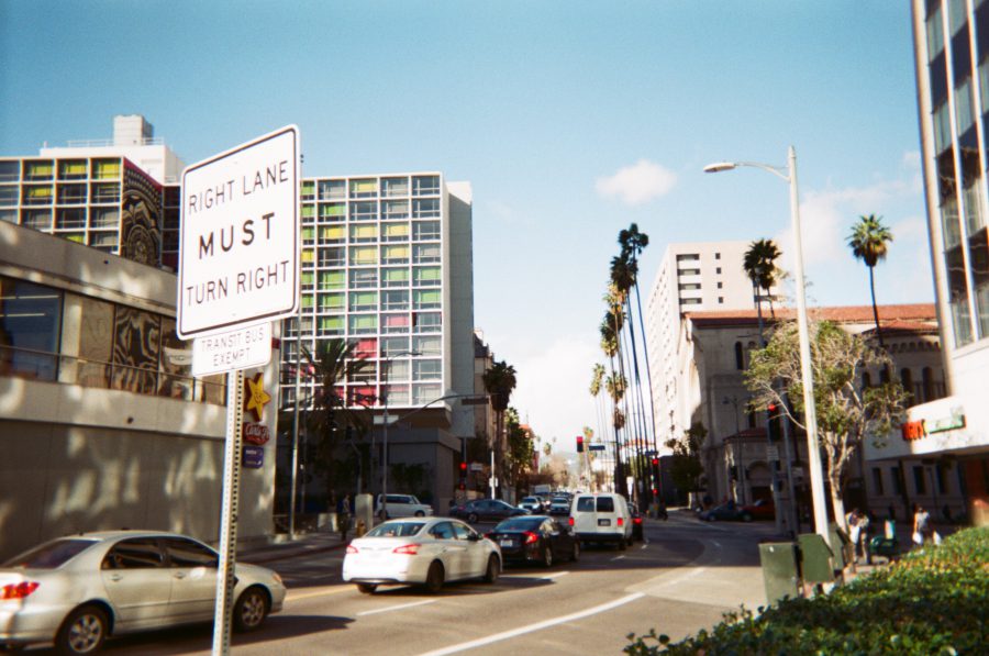 A bustling intersection in West Hollywood lined with tall palm trees, mid-rise buildings, and colorful urban architecture. Cars fill the road under a clear blue sky, and a prominent traffic sign reads "Right Lane Must Turn Right."