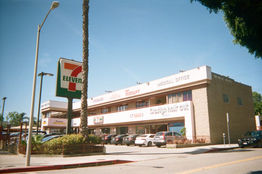 A pink strip mall in West Hollywood under a bright blue sky, with a prominent 7-Eleven sign at the forefront. The building houses a mix of businesses, including a nail salon and a therapy center, Karina's dry cleaners, and the old location of Ben Artikov income tax, with a row of parked cars completing the scene.