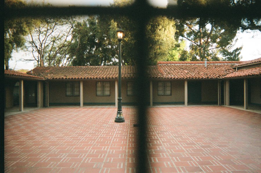 A red-tiled courtyard with a single black streetlamp in the center, viewed through a wrought iron fence. The surrounding buildings have a warm, rustic style with clay-tiled roofs and muted beige walls.
