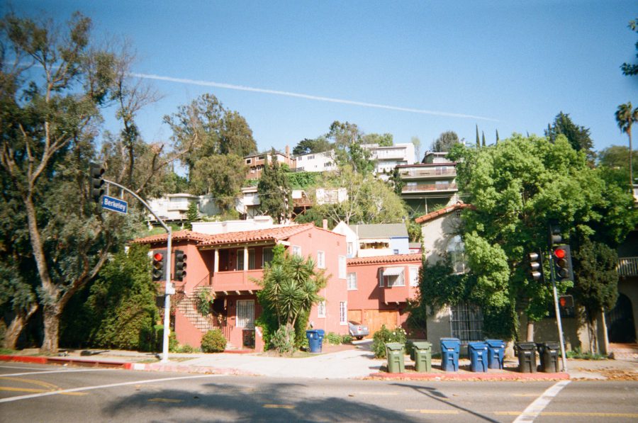 A cluster of homes in a hilly neighborhood near Berkeley Avenue, featuring a vibrant terracotta house with Mediterranean-style architecture in the foreground surrounded by palm trees and greenery, trash and recycle and compost bins lined up neatly along the curb, and additional houses ascending the hillside in the distance.