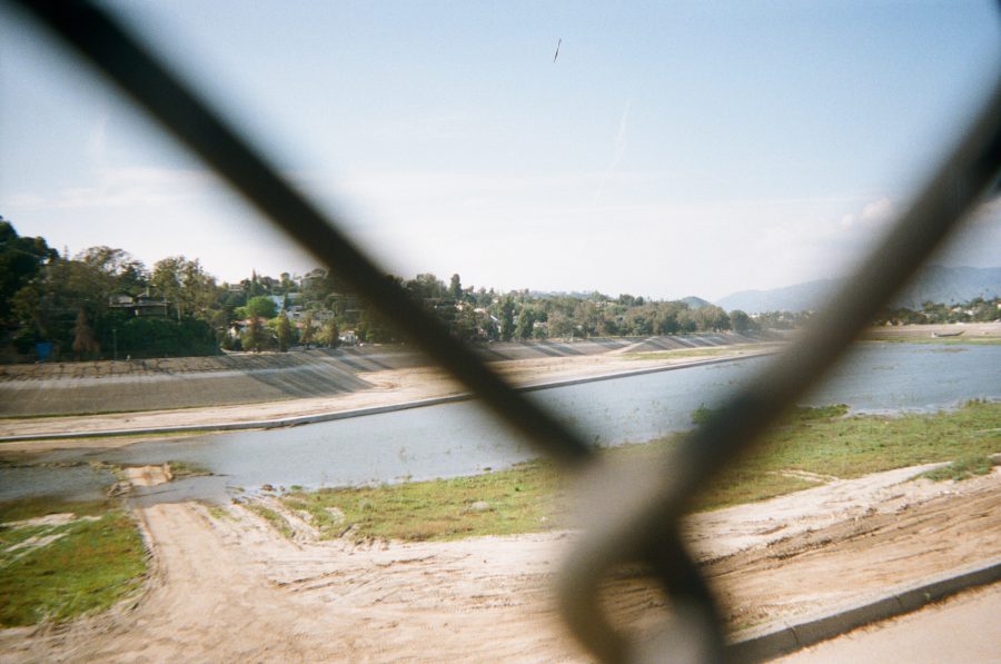 The Silverlake Reservoir seen through a chain-link fence, showcasing a semi-dry waterbed with visible concrete embankments, sparse water, and tree-lined residential areas in the background under a clear blue sky. Silver Lake Reservoir Los Angeles 2017 water level on disposable camera.