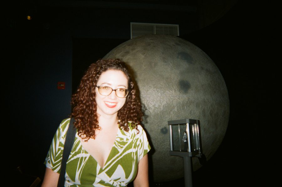 Marina standing in front of a realistic moon installation inside Griffith Observatory. Her curly hair, glasses, and a green and white dress add a touch of elegance. The moon's detailed craters and a nearby vintage telescope create a striking visual contrast.