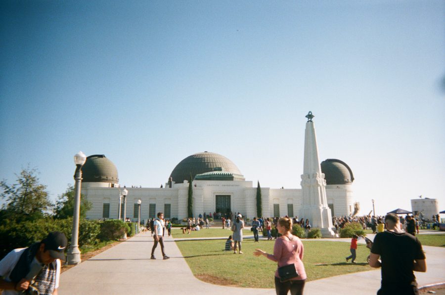 A wide-angle view of the Griffith Observatory, its white walls and iconic domes standing out under a bright blue sky. The foreground is filled with visitors walking through the grassy lawn, with the towering obelisk monument framing the scene on the right.