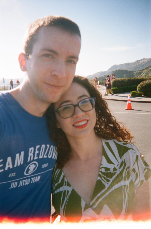 I stand beside Marina at Griffith Observatory, leaning slightly into the frame for a selfie. Marina’s bright red lipstick pops against her curly auburn hair, framing her face, and she wears a green-and-white patterned dress. I wear a blue Team Redzovic Brazilian Jiu Jitsu Team t-shirt, my face turned slightly toward Marina. Along the bottom of the photo, a subtle light leak burn transitions from an orange glow to a white streak, adding a vintage, textured effect to the otherwise crisp and sunny shot.