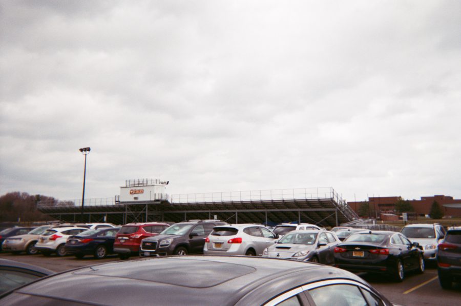 A parking lot on the Rochester Institute of Technology campus filled with various cars under a cloudy sky. In the background, a set of empty metal bleachers marked with the RIT logo stands prominently.
