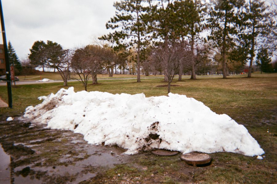 A melting pile of snow in a muddy patch of grass on the RIT campus, surrounded by bare trees and scattered puddles. The background features additional trees and open spaces on a cloudy winter day.