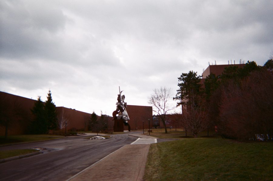 A pathway on the RIT campus leading to a large abstract metal sculpture surrounded by brown-brick academic buildings. Trees and grass frame the area under a cloudy sky.