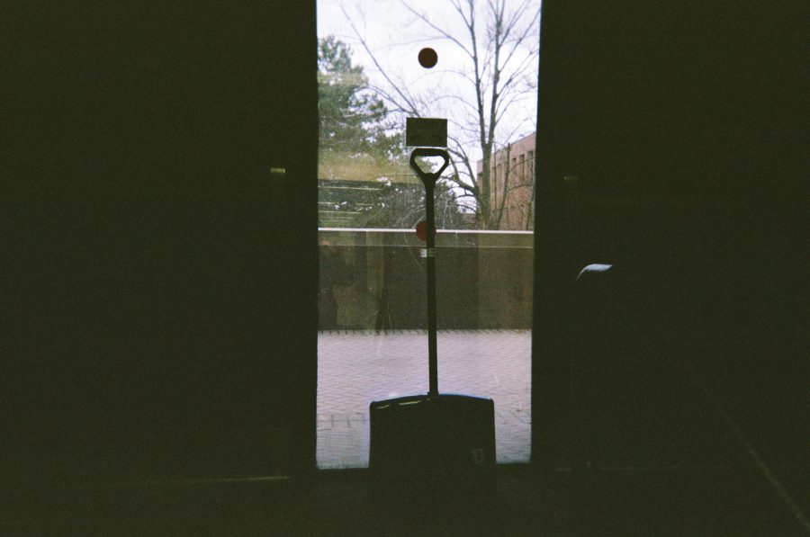A snow shovel propped against the glass doors of an RIT building, viewed from the interior. Beyond the doors, bare trees and paved walkways create a muted outdoor scene.