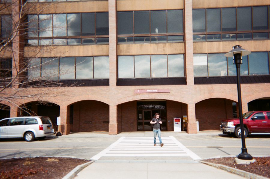 The front of a modern library building at RIT, featuring large glass windows and brick archways. A person walks across the crosswalk in front of the library, with cars parked on either side of the entrance.
