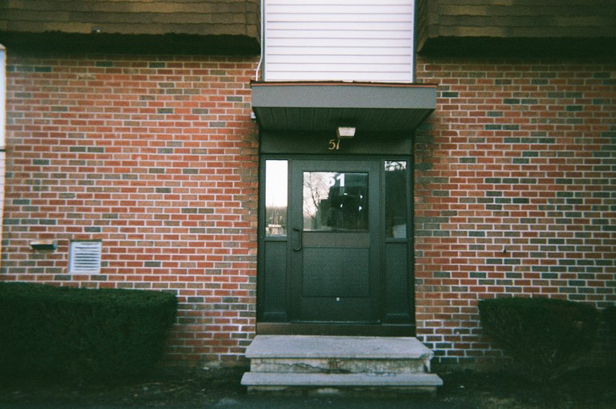 The entryway to an apartment building with a brick façade, black-framed glass door, and number 51 above the door. Trimmed bushes flank the concrete steps leading to the entrance.
