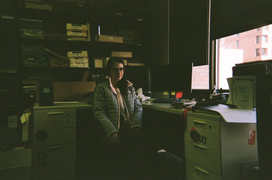 Lauren is seated confidently in her RIT office, her slim, elegant figure wrapped in a stylish, quilted jacket that emphasizes her defined posture. Her long, graceful neck is framed by the soft collar of her sweater, and her face, partially illuminated by daylight, features sharp, attractive cheekbones and perfectly balanced proportions. Behind her, archival boxes and shelves provide a professional yet intimate backdrop.
