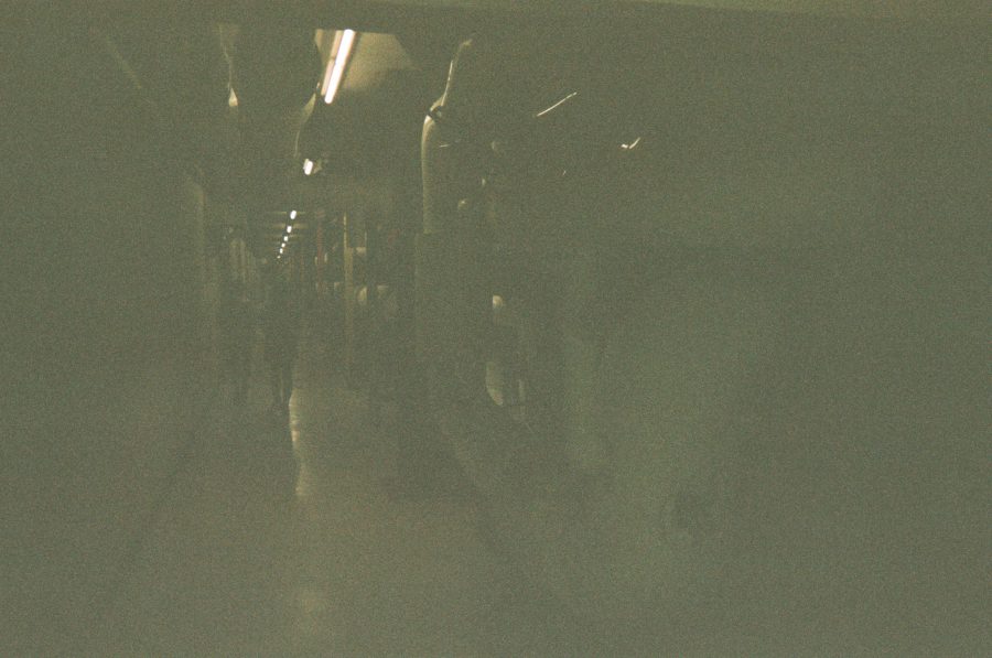 A dimly lit underground tunnel at RIT with fluorescent lights lining the ceiling. Large pipes run along the walls, and faint silhouettes of people are visible in the distance, adding to the industrial atmosphere.