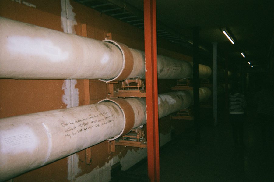 A row of large, industrial pipes attached to the walls in the RIT tunnels. One pipe is marked with more graffiti, including humorous and cryptic messages, set against the backdrop of worn orange and white walls.