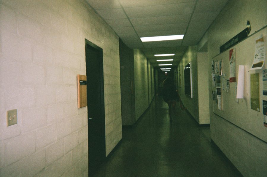 Sam walking down a long, fluorescent-lit hallway at RIT, her figure slightly blurred in motion. The cinderblock walls and bulletin board with flyers add to the institutional feel of the space.