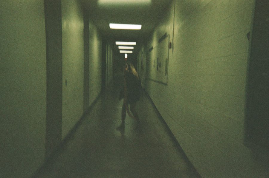 Sam is captured mid-run in a fluorescent-lit hallway at RIT. The hallway’s white cinderblock walls are lined with bulletin boards, and her motion blurs slightly, adding dynamic energy to the image.