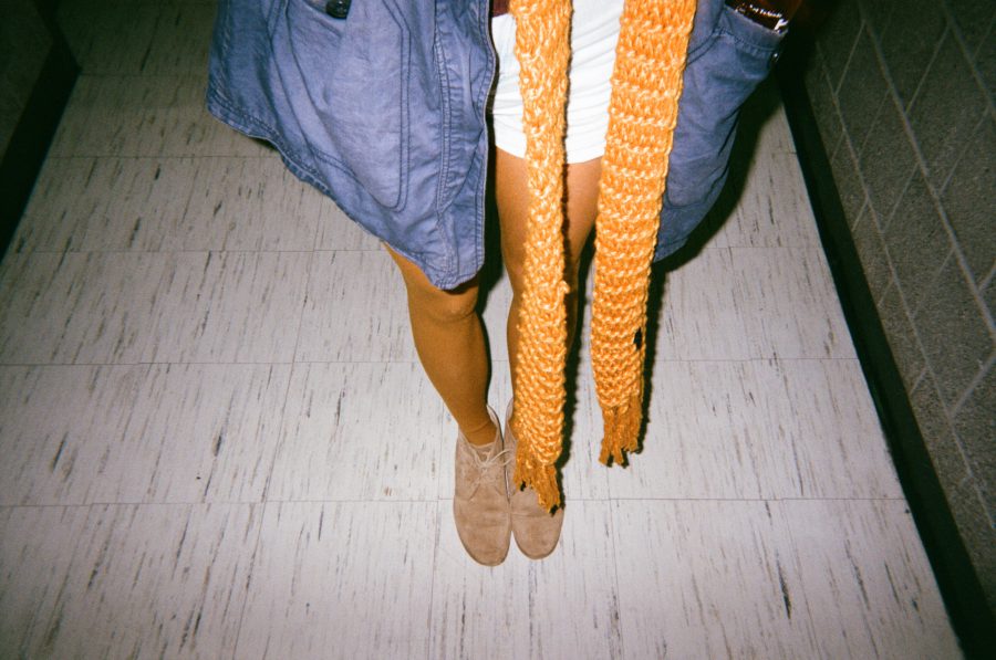 A close-up of Sam’s lower half as she refuses to have a face photo in the RIT hallway. She wears a chambray jacket over white shorts with an orange knit scarf and beige ankle boots. The polished tile floor reflects the soft indoor lighting.
