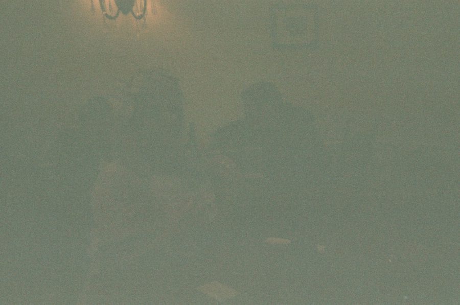 Lauren sitting in front of a glowing candlelit table, her figure barely visible through the grainy disposable film. The low light creates a warm ambiance, with soft shadows blending into the muted background.