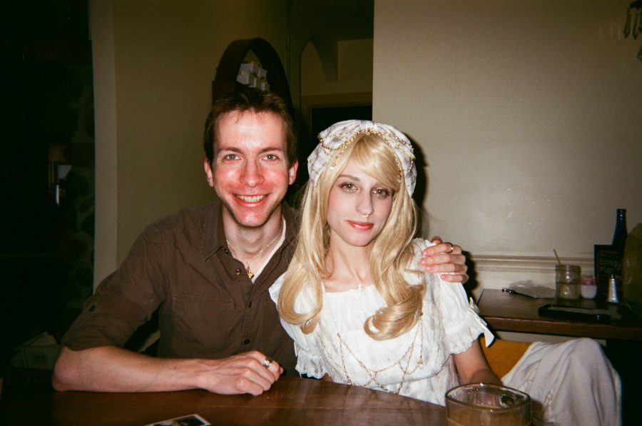 At the restaurant, Lauren sits beside me, wearing an exquisite white gothic lolita dress that hugs her slender waist and cascades into a delicately embroidered skirt. The fitted bodice showcases her refined figure, while her golden blonde wig, styled in soft curls, frames her glowing complexion. The intricate lace detailing and pearl accessories of her outfit only add to the sophisticated allure of her radiant presence.