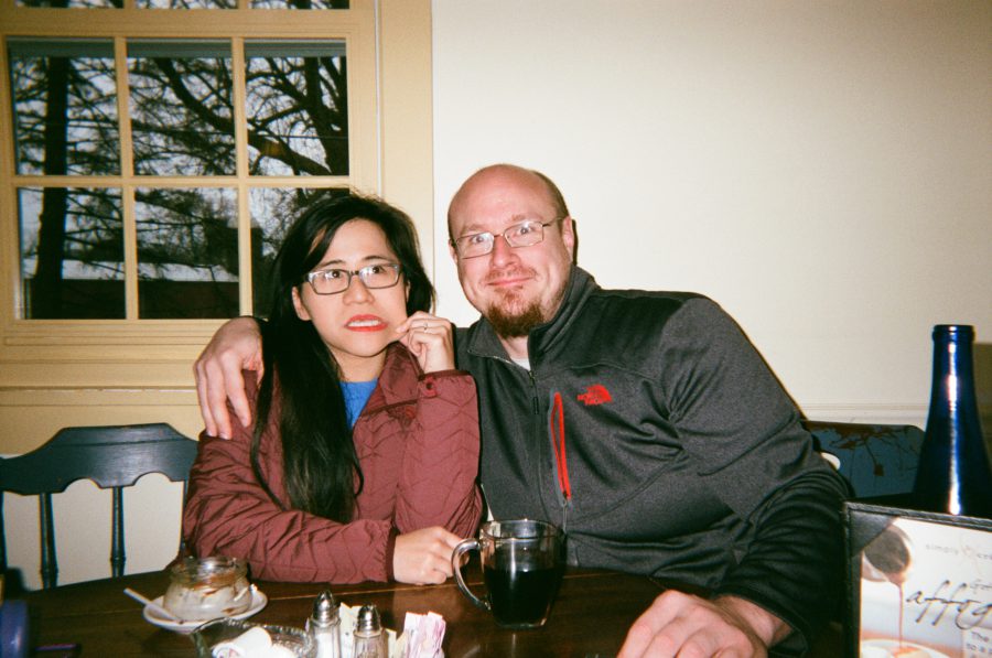 Lauren’s two friends seated at the same restaurant. One wears a red jacket and glasses, posing with a thoughtful expression. The other has on a gray zip-up sweater, smiling broadly, as a window with bare winter trees provides the background.