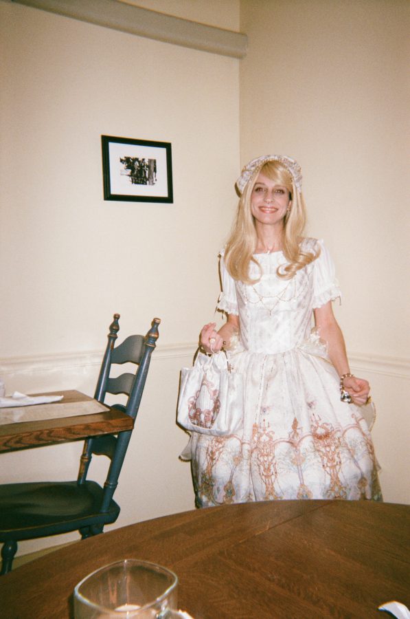 Lauren stands beside the table in the restaurant, her white gothic lolita dress perfectly complementing her petite frame and graceful posture. The fitted design emphasizes her narrow waist, while the intricate pastel designs on the fabric draw attention to the flowing hemline of her skirt. Her elegant hands rest gently on a vintage-inspired purse, adding a sense of refined charm to her striking silhouette.
