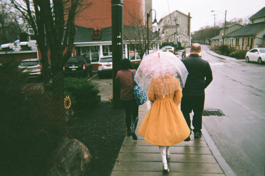 Lauren walking outside under an umbrella with a clear canopy decorated with pink floral designs. She wears a bright yellow coat, contrasting against the wet sidewalk and overcast Rochester streetscape with houses and cars in the background. Her two friends walk in front of her.