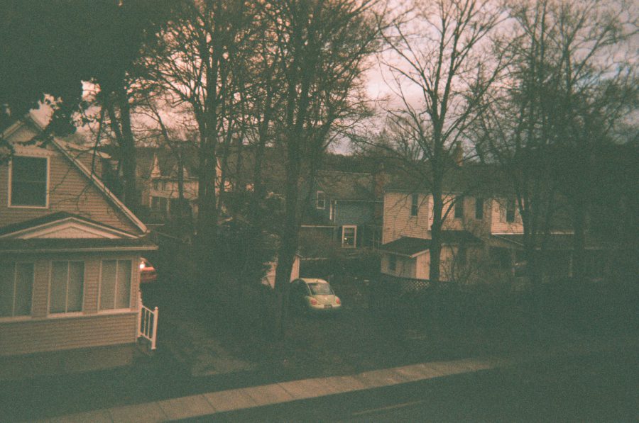 A view from my Airbnb window overlooking a residential Rochester neighborhood. The scene includes a cluster of older houses surrounded by bare trees, with a green Volkswagen Beetle parked in a backyard driveway.