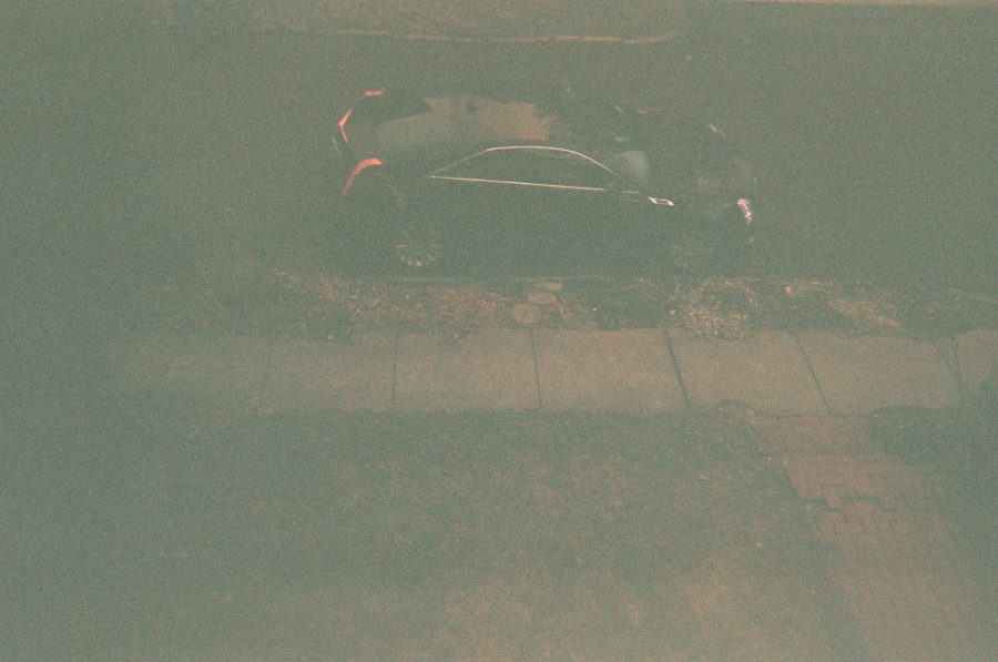 A nighttime view from my Airbnb window, capturing the dimly lit and rainy sidewalk and street below. A dark car is parked along the curb, its taillights glowing faintly under the streetlamp.