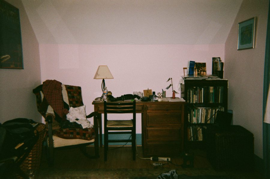 A cozy Airbnb bedroom with a small wooden desk, a chair, and a tall bookshelf filled with books on the right. A colorful patchwork blanket drapes over a chair to the left, and the space is softly lit by a table lamp on the desk.