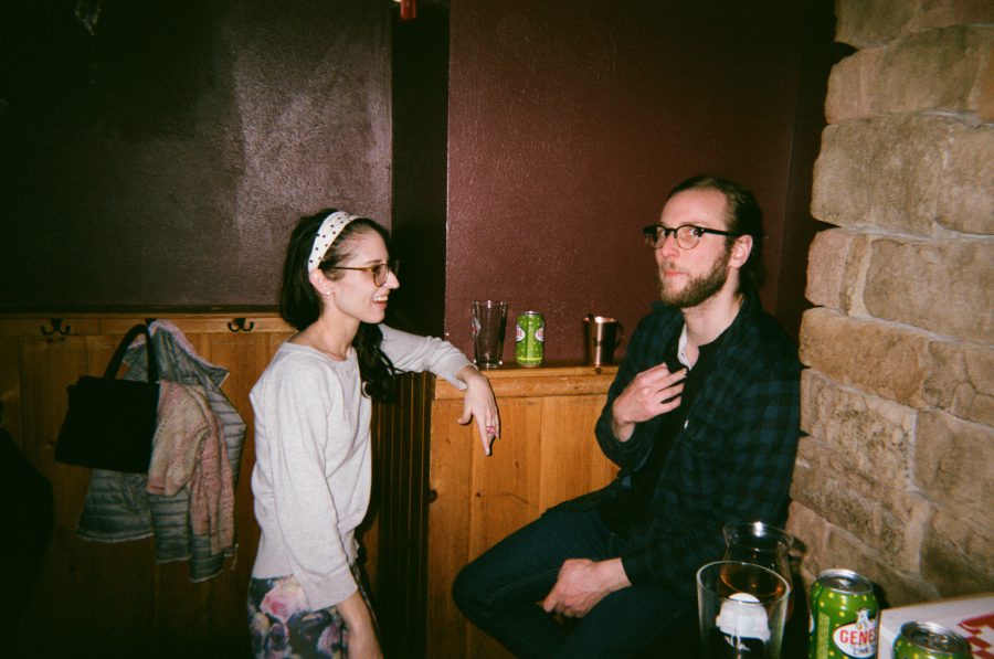 At a Rochester bar, Lauren stands on the left, wearing glasses and a white headband, leaning against a wooden divider while talking to a man with glasses and a plaid shirt seated next to her. Drinks and soda cans sit on a small ledge nearby.