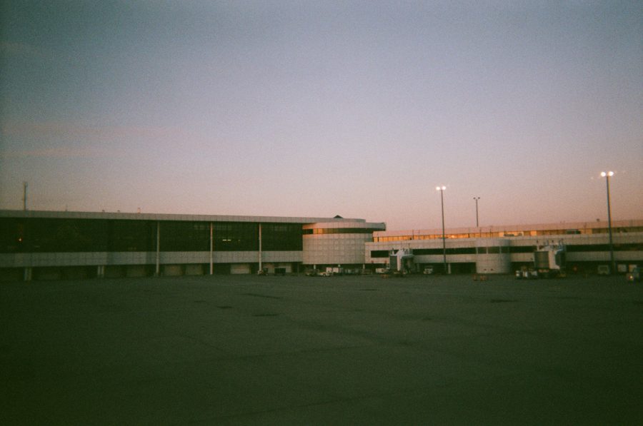 The exterior of Rochester airport at dusk, with the large terminal building stretching across the image, bathed in a soft orange glow from the setting sun. Airport equipment and vehicles are visible near the building.