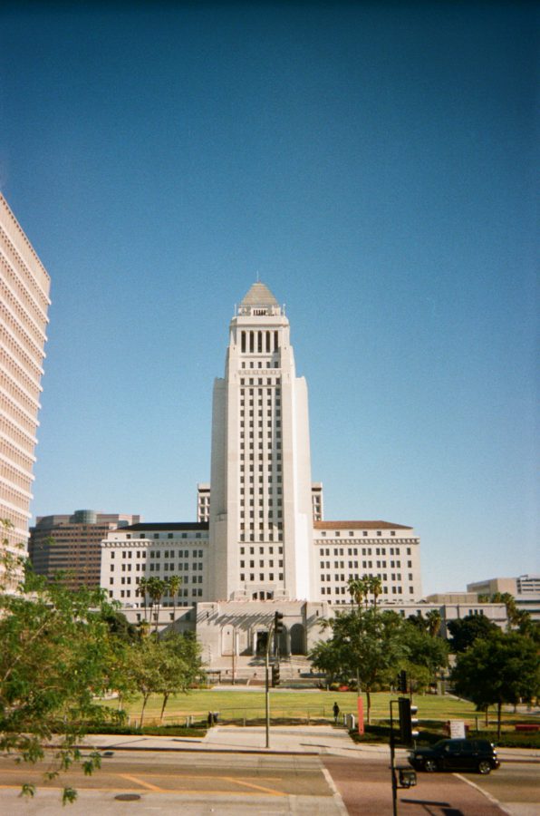 Los Angeles City Hall captured against a clear blue sky. The towering white building features intricate art deco architecture, flanked by lush green trees in Grand Park. Surrounding buildings frame the shot, emphasizing the iconic structure's prominence.
