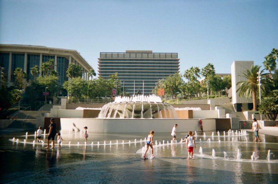 A wide view of the fountains at Grand Park in DTLA. Jets of water arc up from the ground, with children and adults playing in the shallow pool below. The backdrop features a tiered fountain, palm trees, and the imposing architecture of the Department of Water and Power building.