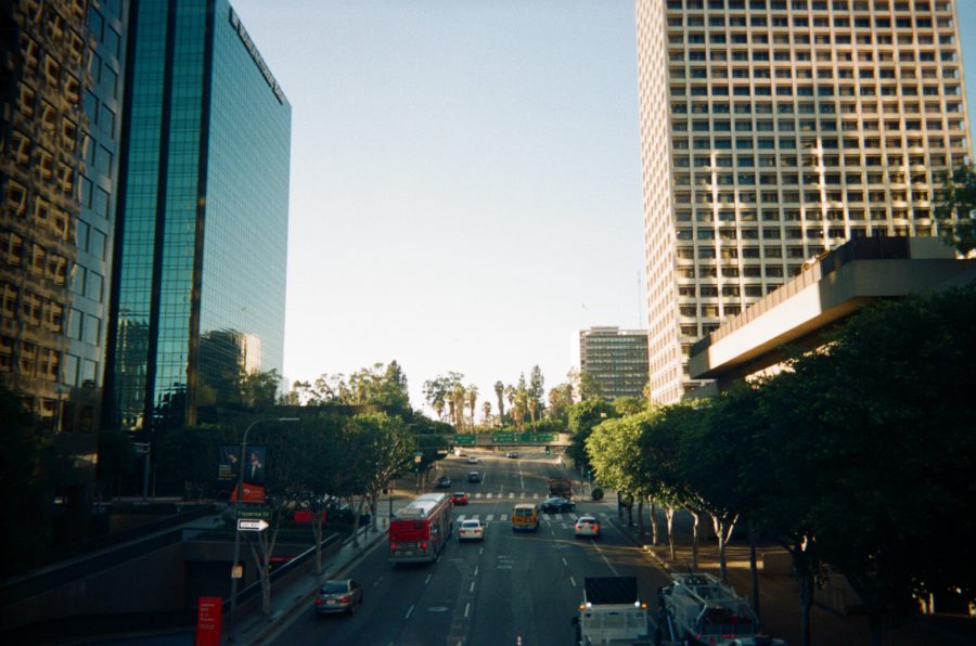 Overlooking a busy Los Angeles street in the financial district. Tall modern office buildings with reflective glass rise on either side of the road, while smaller trees line the sidewalks. Cars and buses navigate the bustling intersection below.