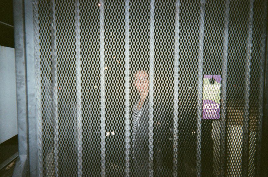 Katie standing behind a grated metal gate door, illuminated by a bright camera flash. Her face is partially visible through the crisscross pattern of the gate. A purple and green notice and dark background contrast against the steel mesh.