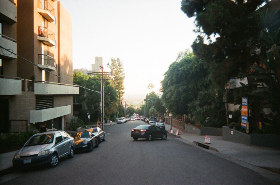 A gently sloping road in Hollywood just south of Runyon Canyon, framed by mid-century apartment buildings and lush greenery. Cars are parked neatly along both sides of the street, and the view extends downhill toward a hazy horizon. Distant palm trees dot the skyline.