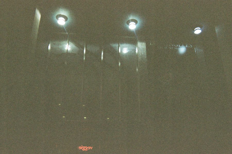 The Los Angeles Film School building stands prominently against a dark backdrop. Spotlights shine down from above, creating reflective streaks on the smooth surface of the building's entrance. The school's name is partially visible in faint lettering, and the muted lighting adds a cinematic mood to the photo.