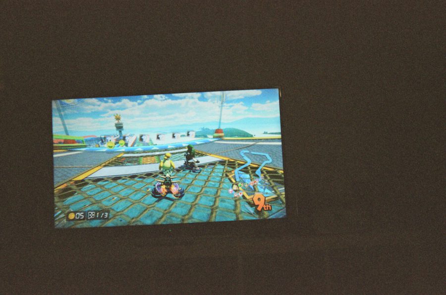 A television screen displays a Mario Kart 8 game in progress. Isabelle, a character from the Animal Crossing series, is the main racer in view, driving a small go-kart on a vibrant track with blue skies and detailed surroundings. The game HUD shows "9th" place, and the colorful graphics contrast against the darkened apartment living room. I'm the one playing; Katie would be in first place.