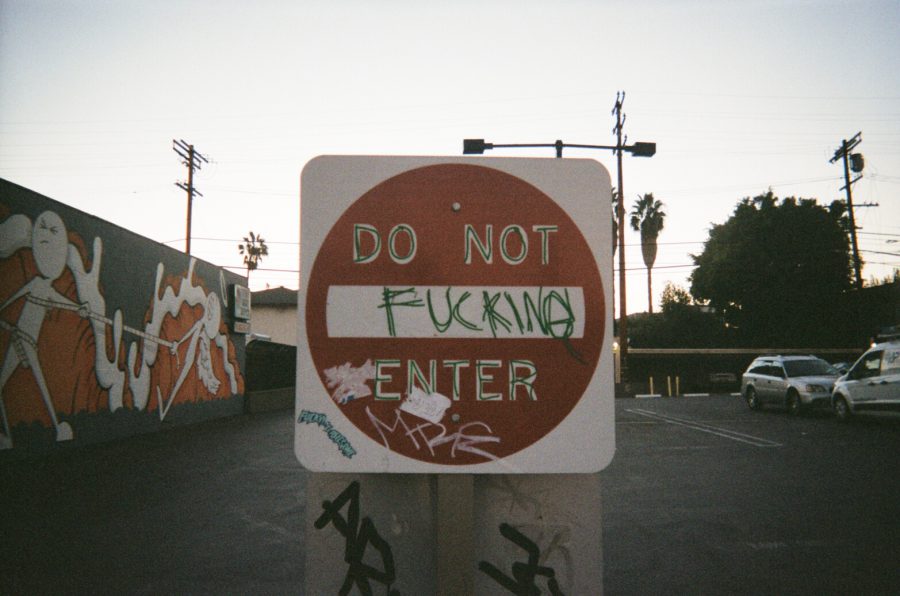 A vandalized "Do Not Enter" sign in a Los Angeles parking lot has been spray-painted to read "Do Not Fucking Enter." To the left, a mural on a building features dynamic orange and white illustrations of abstract humanoid figures with elongated limbs and bold outlines. The street is framed by palm trees and utility poles in the background, capturing a gritty yet artistic LA vibe.
