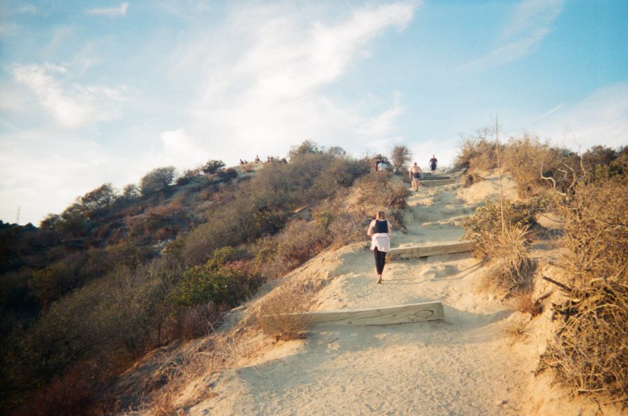 A hiking trail in Runyon Canyon with steps reinforced by wooden beams to combat erosion. The path climbs a dry hillside dotted with sparse shrubs and bushes. Several people hike up the trail, some wearing athletic clothing and carrying water bottles. The hilltop is faintly visible at the top of the frame, with a few hikers already at the summit.
