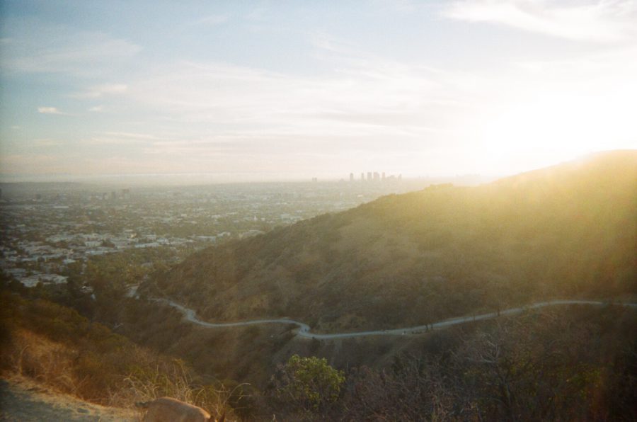 A view of Runyon Canyon during sunset. The trail winds through the hills, with dry vegetation on either side. The light creates a warm glow over the landscape, while the horizon shows a distant city skyline, likely Century City, and a faint glimpse of the Pacific Ocean further out. The golden hour lighting adds depth to the natural and urban scenery.