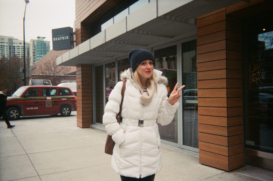 My friend Ashley posing in front of Beatrix coffee shop in Chicago. She is dressed warmly in a belted white winter coat with a faux-fur hood, a beige knit scarf, and a dark gray beanie. Her blonde hair peeks out beneath the hat, and she’s holding up a peace sign with her right hand. Ashley’s bright red lipstick stands out against her light complexion. The coffee shop has a modern wooden facade, and a red taxi is parked on the street behind her.