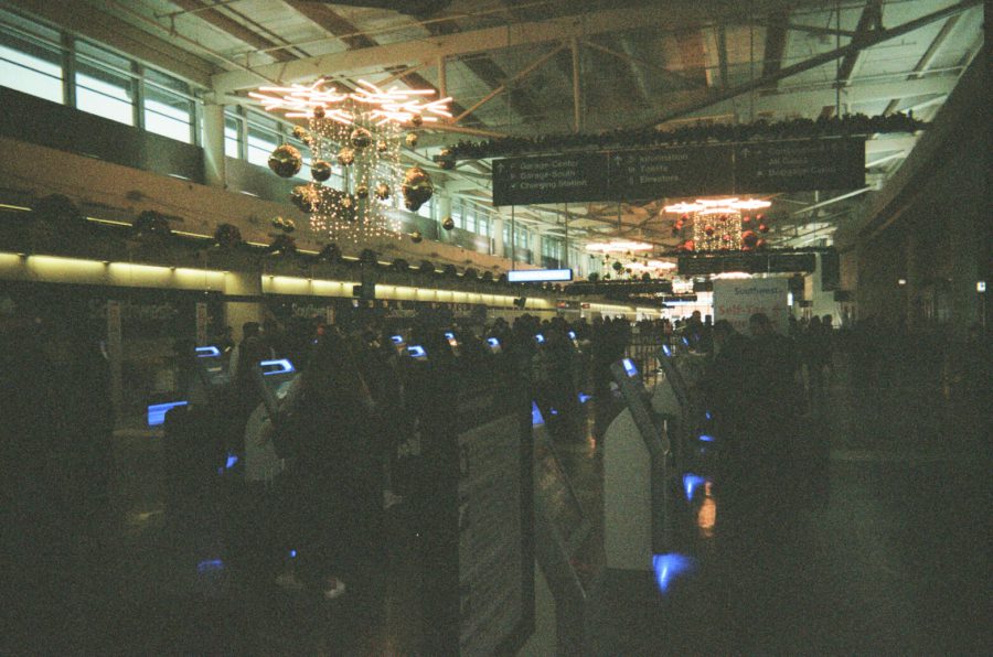 An interior shot of an airport terminal in Chicago, decorated for the holiday season. The ceiling is adorned with illuminated ornaments, including starburst-like designs and hanging baubles. Check-in kiosks glow with blue light, and travelers are seen milling around the terminal. Overhead signs display flight information, and the scene captures the bustling yet festive atmosphere of holiday travel.