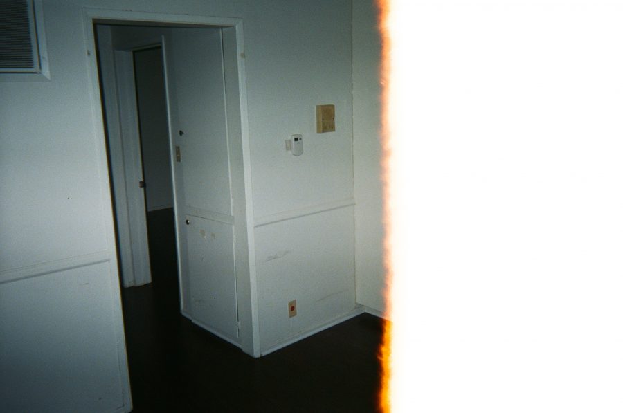 A final image with a dramatic light leak on the right, casting a bright orange and white streak over the photo. The view shows the hallway of the apartment, with white walls and dark brown hardwood floors. A door to the bedroom is open, and the faint outlines of wall-mounted controls and light switches are visible on the left.