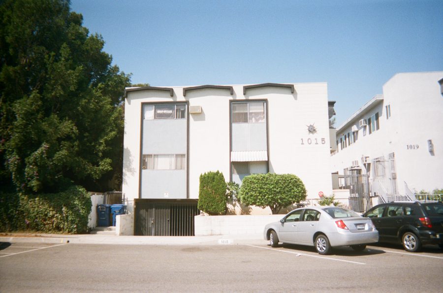 The front façade of 1015 N Vista Street, viewed from across the road. The building is painted white with light gray accents, featuring two levels of windows. There are trash bins to the side, a sloped driveway down to below the building to the gated garage, and shrubs in front of the rest of the building. A few parked cars, including a silver sedan, are in the foreground, and tall green trees frame the scene on the left.