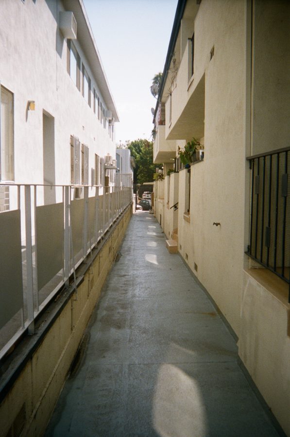 A long, narrow paved side pathway between two buildings, leading to the front gate and the street. The walls of the buildings are painted light beige, and small windows face the path. The ground is smooth concrete, with patches of light and shadow created by the afternoon sun. A glimpse of the street and parked cars is visible at the far end.
