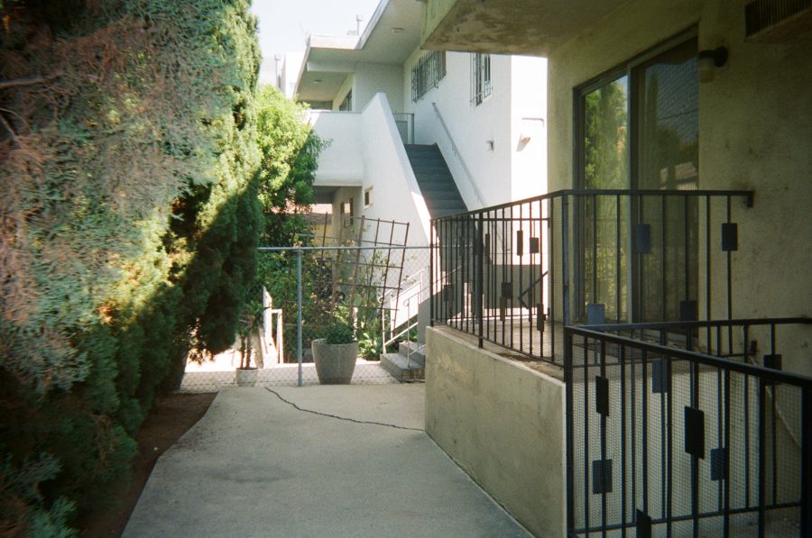 An outdoor paved area behind the apartment building, showing a raised, fenced-in patio area surrounded by a black metal railing. Neighboring stairs and a second-floor landing are visible beyond the patio. To the left, tall green trees provide shade, while a few potted plants sit on the concrete surface.