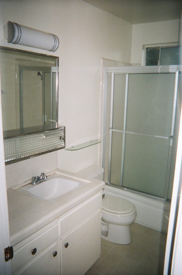 A compact bathroom with white tiled walls and a sliding glass shower door. The sink is set into a white vanity with a chrome faucet and silver knobs. Above the sink, a mirrored medicine cabinet reflects the white walls. A small glass shelf is mounted to the right, and the toilet is positioned beside the shower. The flooring is cream-colored linoleum.