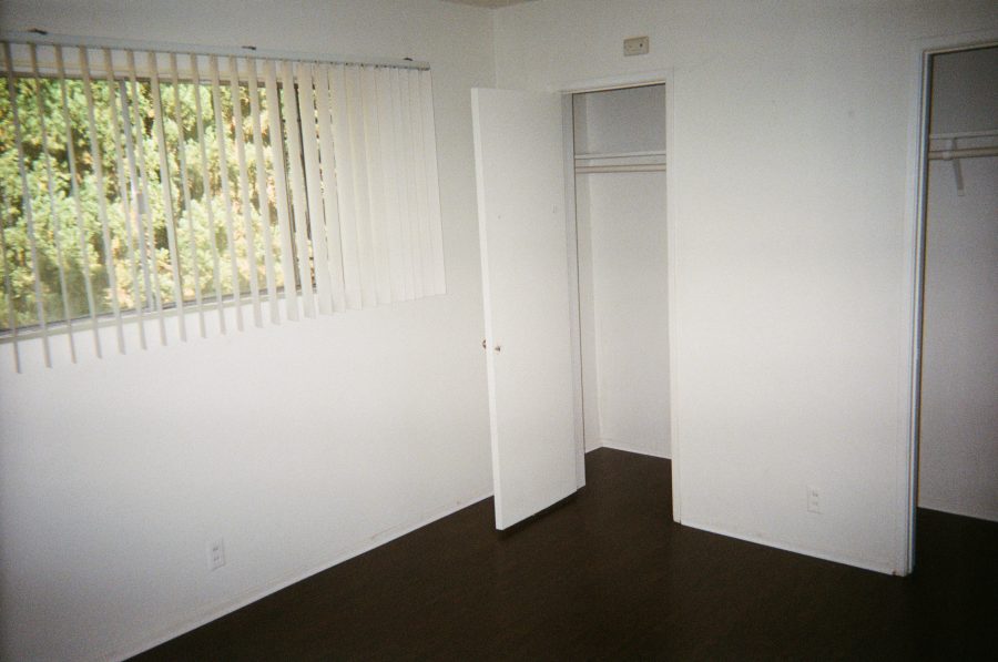 The bedroom in our West Hollywood apartment, now empty. The room has dark brown hardwood floors, a large window with vertical blinds partially drawn revealing foliage outside, and a double closet with both doors open. Inside the closet are a single wooden shelf and hanging rail. The walls are painted white.